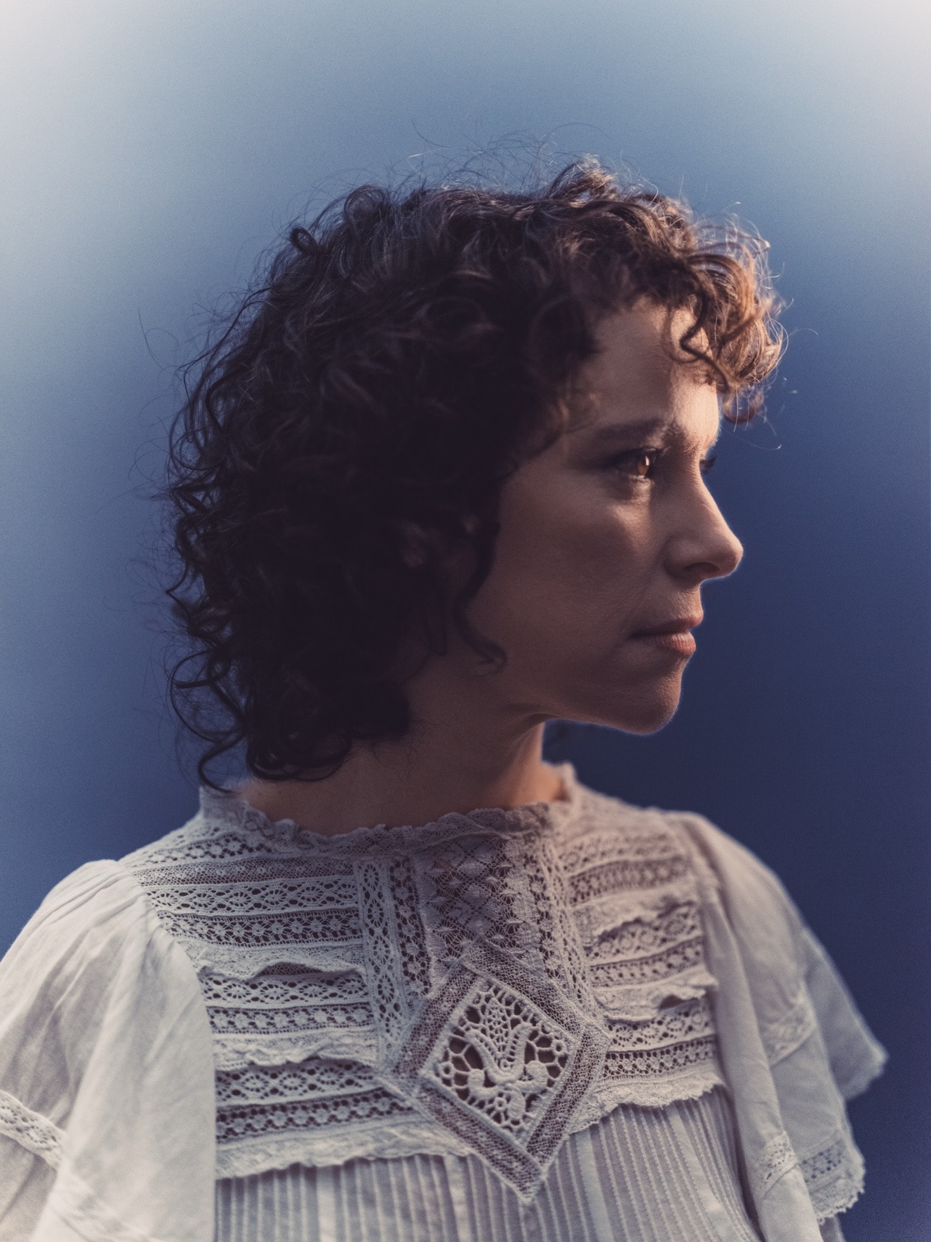 Emily Loizeau de profil sur fond bleu, chemisier en dentelle claire.