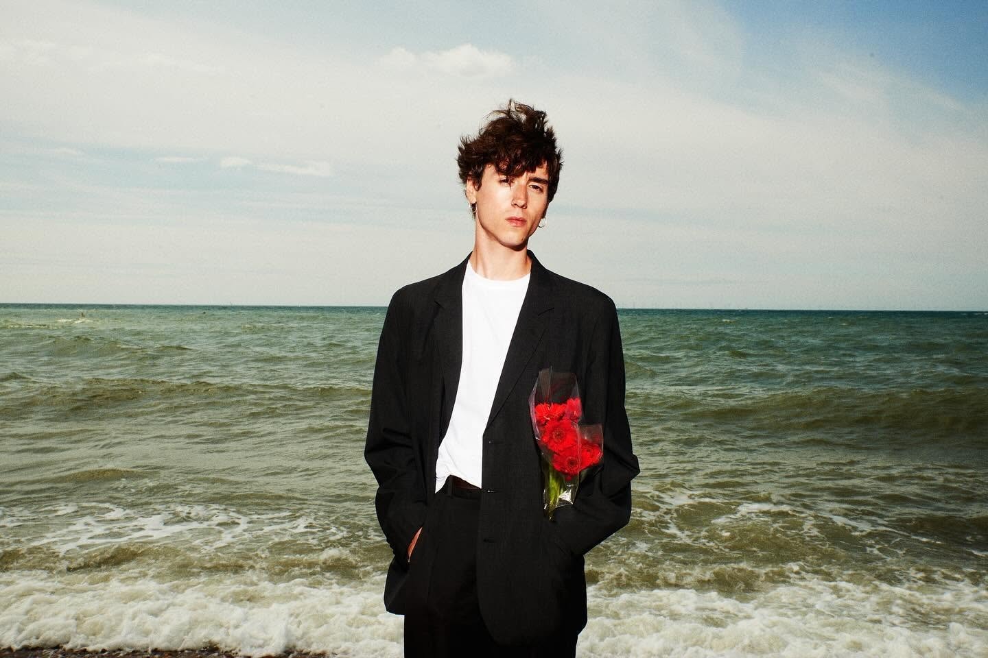 Abel en costume noir devant la mer, un bouquet de roses rouges à la main.