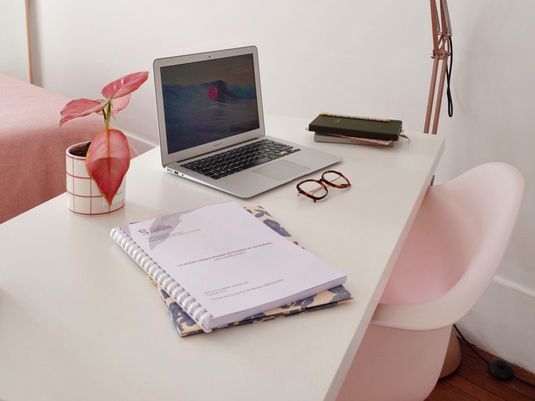 Un bureau blanc : un ordinateur portable ouvert, une plante aux feuilles roses, une paire de lunettes et un carnet à spirale.